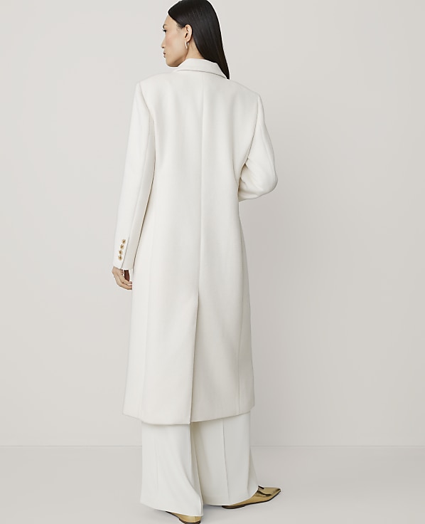 A woman in a long white coat and wide-leg pants stands against a plain, light background.