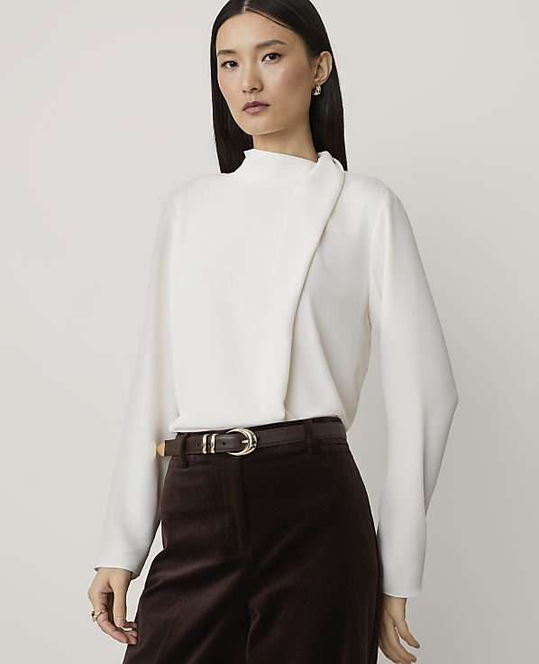 A woman in a white blouse and brown pants stands against a plain background.