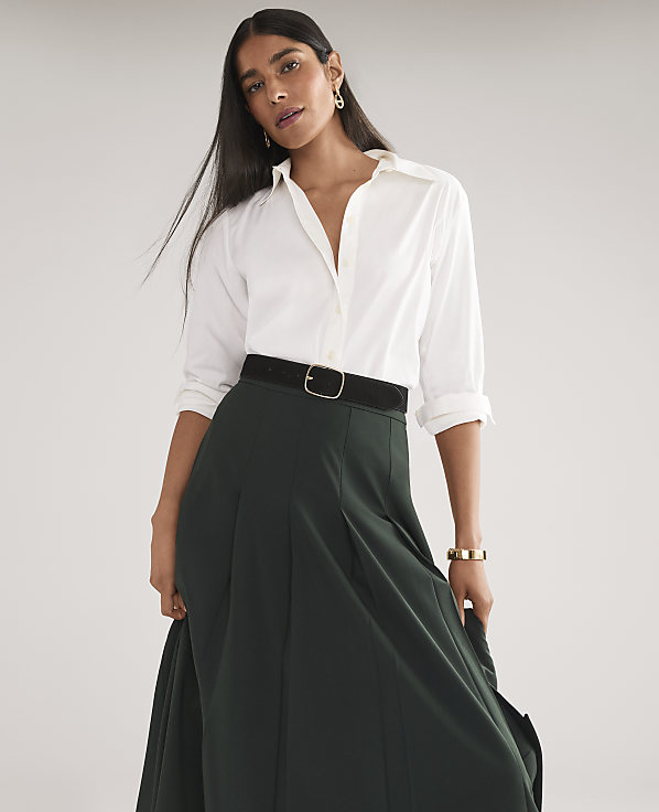 A woman in a white shirt and dark green skirt stands against a plain, light gray background.