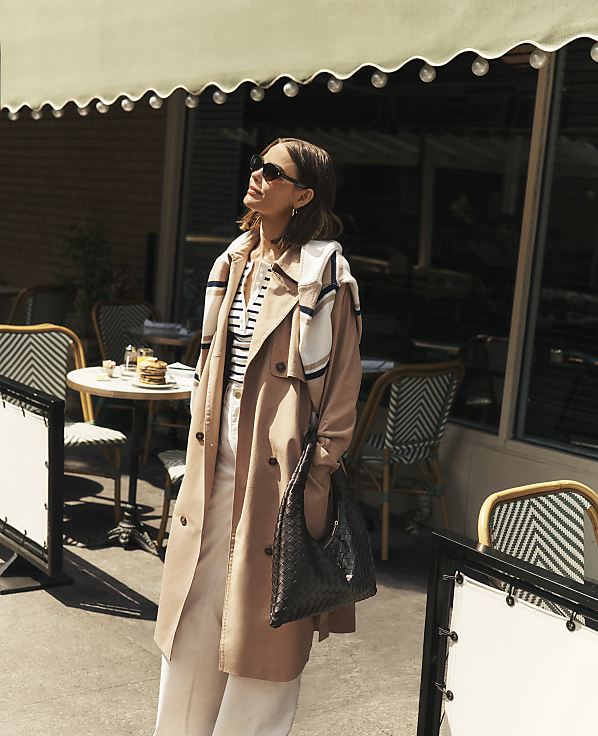 A woman in a trench coat stands in front of a cafe with tables and chairs visible in the background.