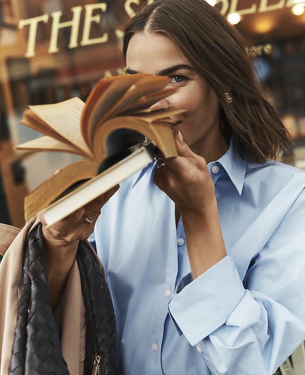 A woman in a blue shirt holds an open book in front of her face, with a blurred storefront in the background.