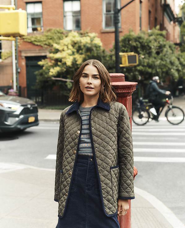 A woman in a quilted jacket stands on a sidewalk with a blurred city street and a cyclist in the background.