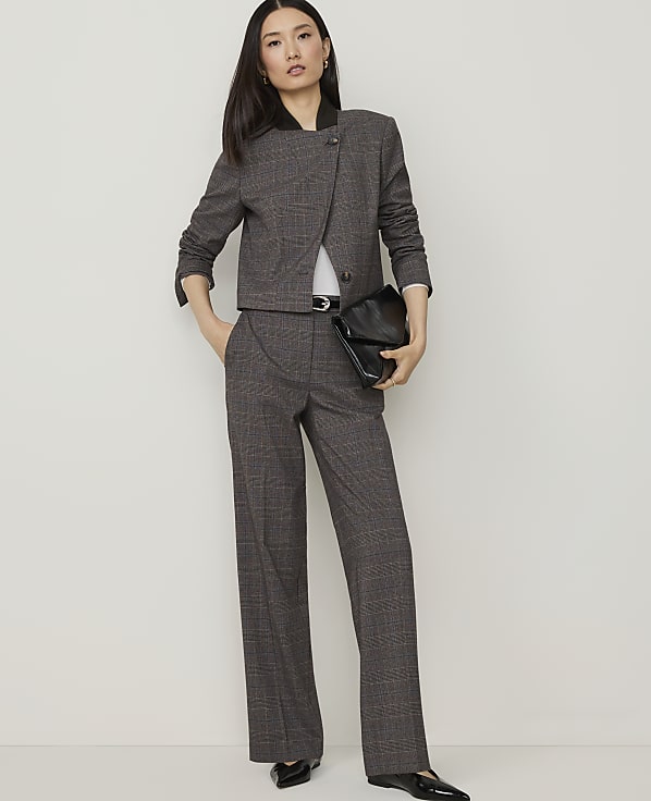 A woman in a black turtleneck and plaid pants stands against a plain white wall, holding a black tote bag.