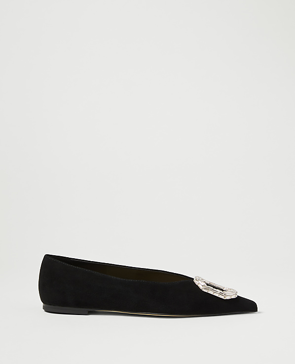 A black suede flat shoe with a jeweled buckle sits on a plain white background.