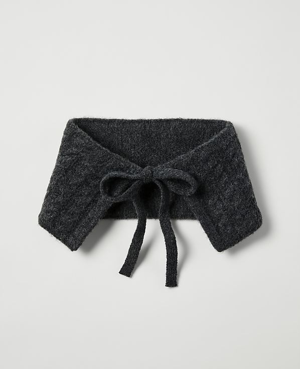 A dark gray knitted collar with a bow in the foreground rests on a plain, light gray background.