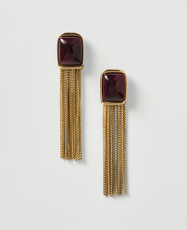 A pair of gold fringe earrings with deep red square gems hang against a plain white background.