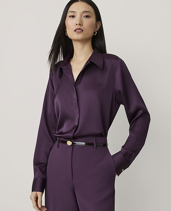 A woman in a purple satin blouse and matching pants stands against a plain, light gray background.