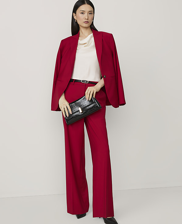 A woman in a red suit and cream top holds a black clutch in front of a plain white background.
