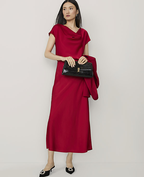 A woman in a red dress and heels stands with a black clutch and jacket against a plain white background.