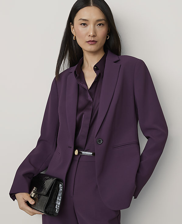 A woman in a purple suit holds a black clutch in front of a plain gray background.