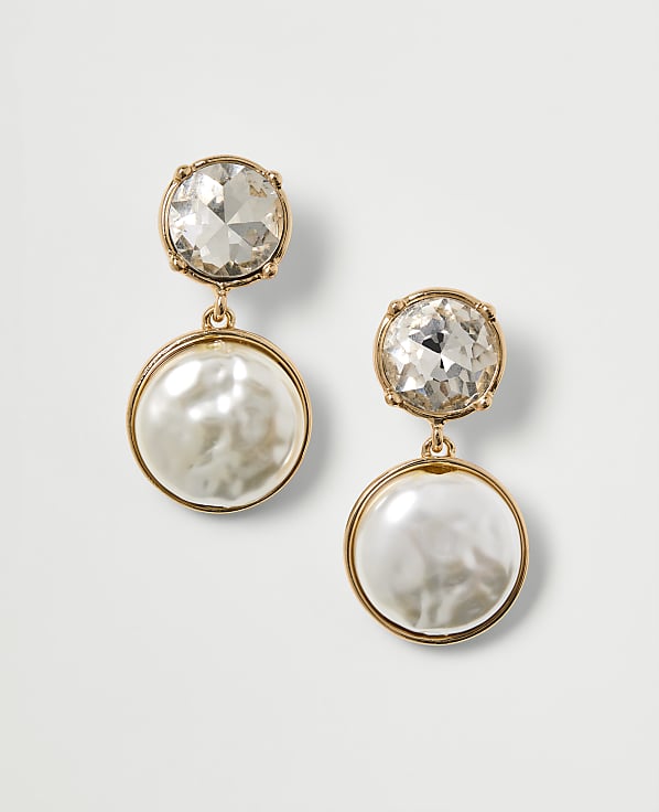 A pair of gold-toned drop earrings, each featuring a clear crystal stud above a lustrous pearl disc, are presented against a plain white background.
