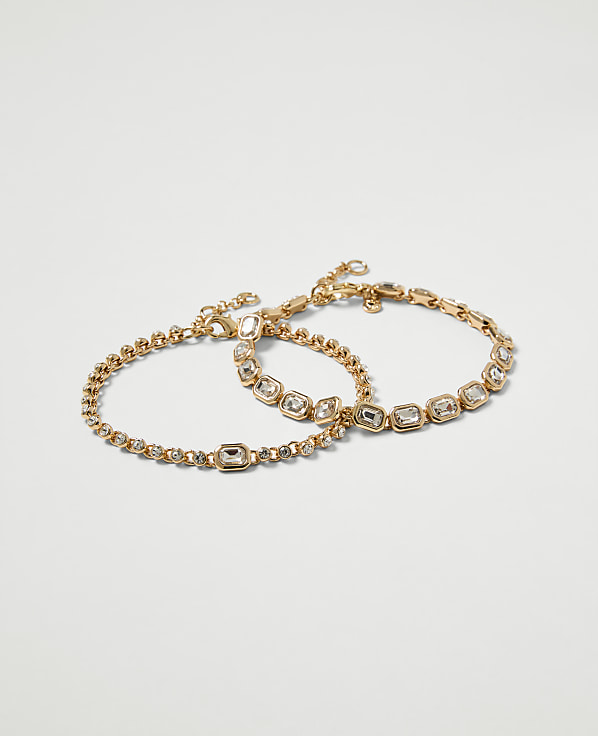 Two delicate gold bracelets, one with a simple chain and the other adorned with clear stones, rest against a plain white background.