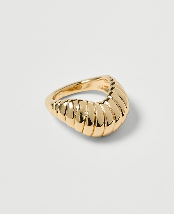 A gold ring with a ribbed, curved design rests on a plain, light gray background.