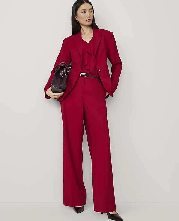 A woman in a red pantsuit stands against a plain white background, holding a dark brown handbag.