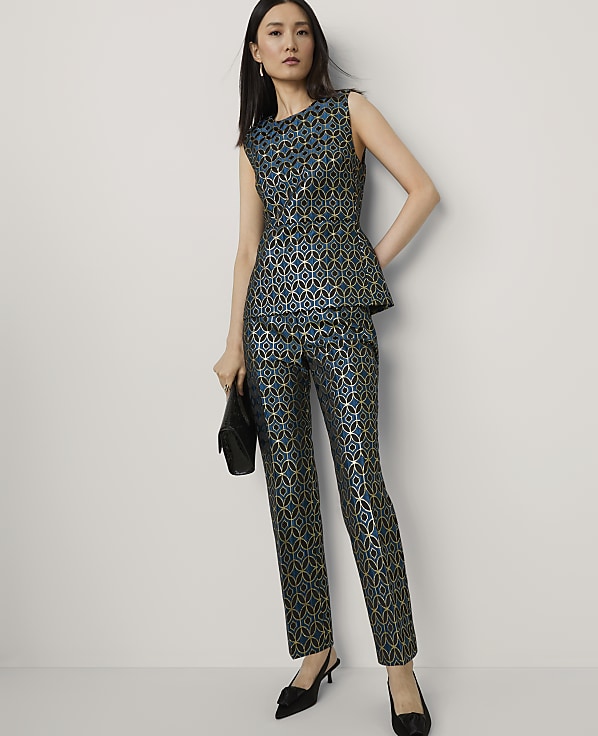 A woman in a teal and gold patterned peplum top and matching pants stands with a black clutch against a plain white background.