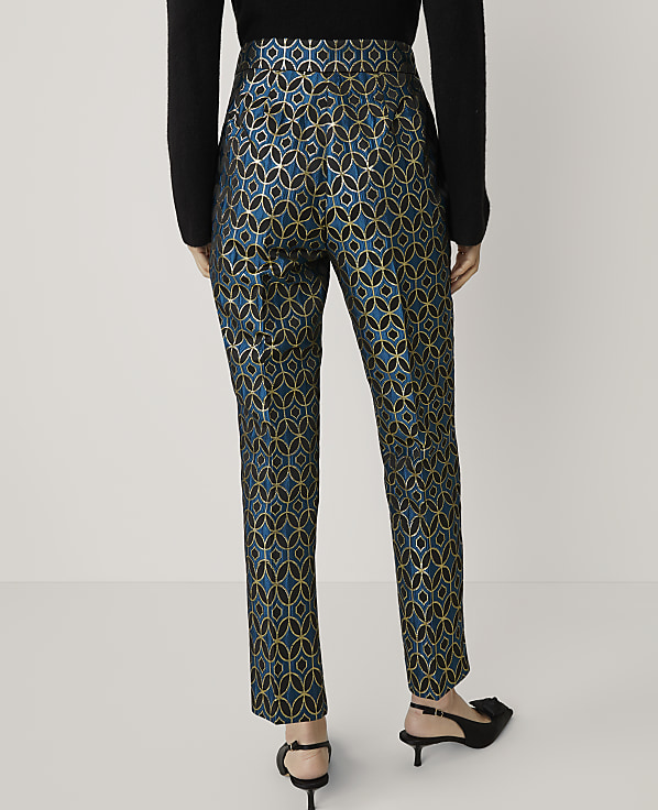 A person stands in front of a plain white background, wearing patterned teal and gold trousers and black heels.