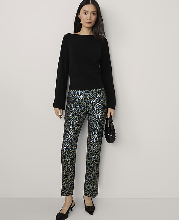 A woman in a black sweater and patterned pants stands against a plain background.