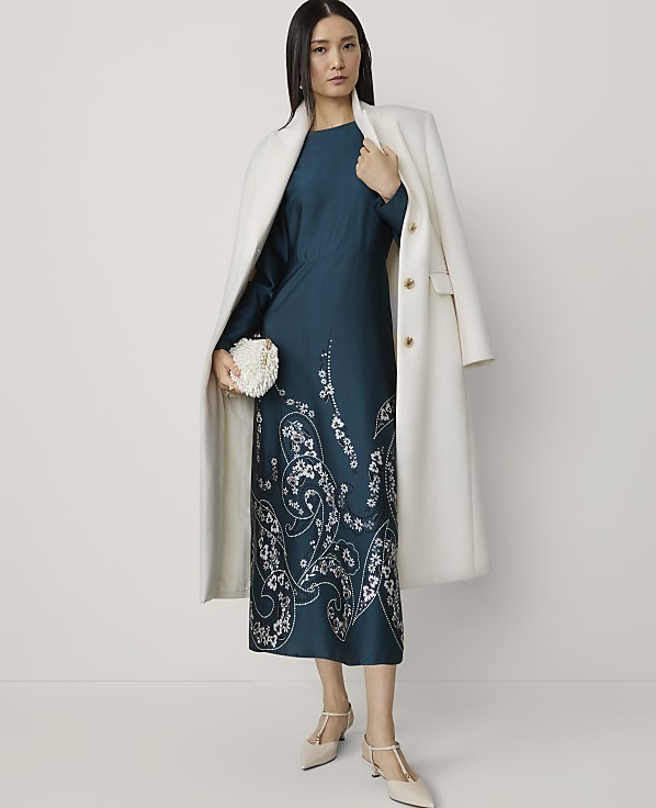 A woman in a teal dress with white floral embroidery and a white coat stands against a plain white background, holding a white clutch.