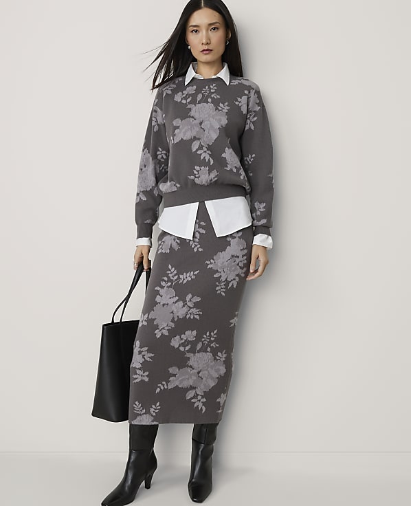 A woman in a floral sweater and skirt stands against a plain background, holding a black tote bag.