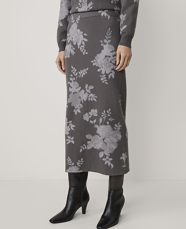 A person stands wearing a floral-patterned skirt and sweater, with black boots, against a plain white background.