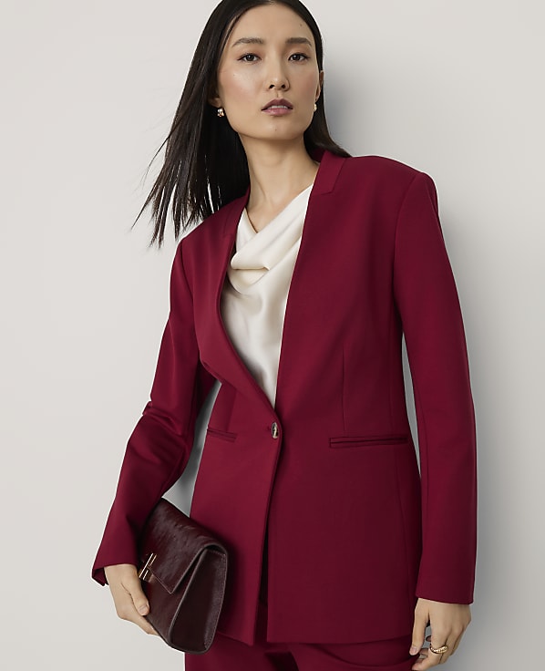 A woman in a burgundy blazer and matching pants holds a clutch bag against a plain background.