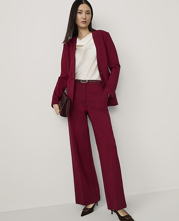 A woman in a burgundy suit and cream blouse stands against a plain white background, holding a burgundy clutch.