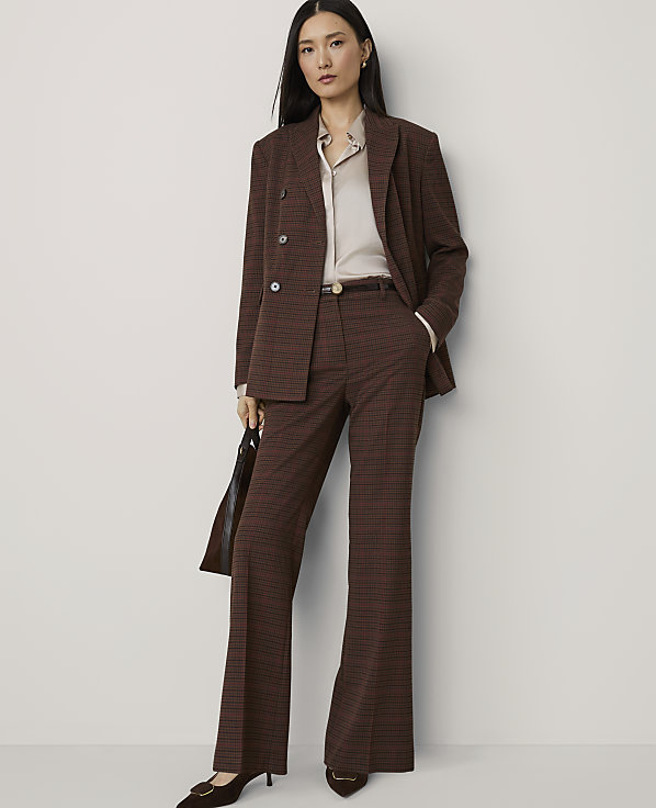 A woman in a brown plaid suit stands against a plain, light-colored background, holding a dark handbag.