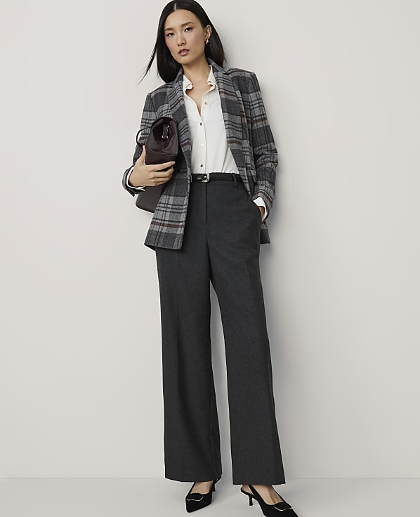 A woman in a plaid blazer and wide-leg pants stands against a plain white background, holding a burgundy handbag.