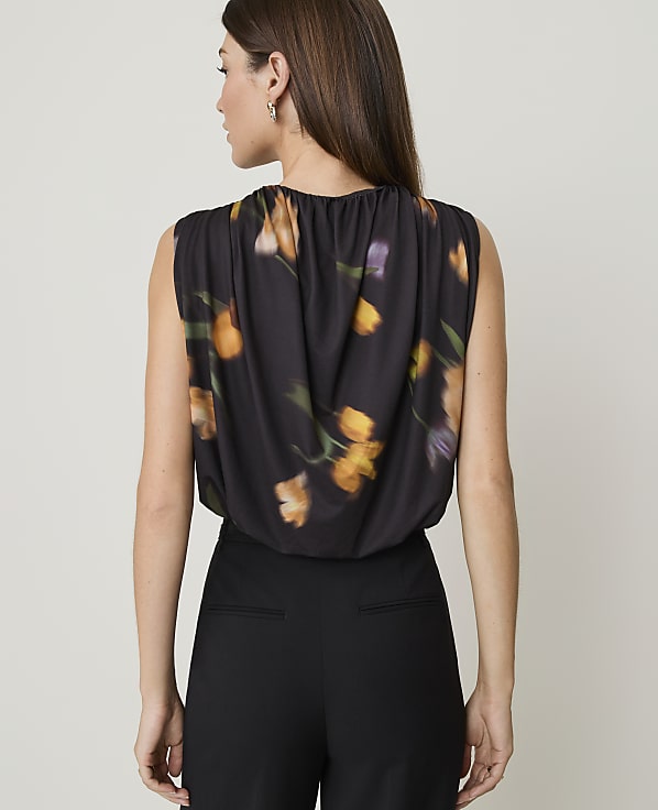 A woman in a floral print top and black pants stands against a plain, light-colored background.