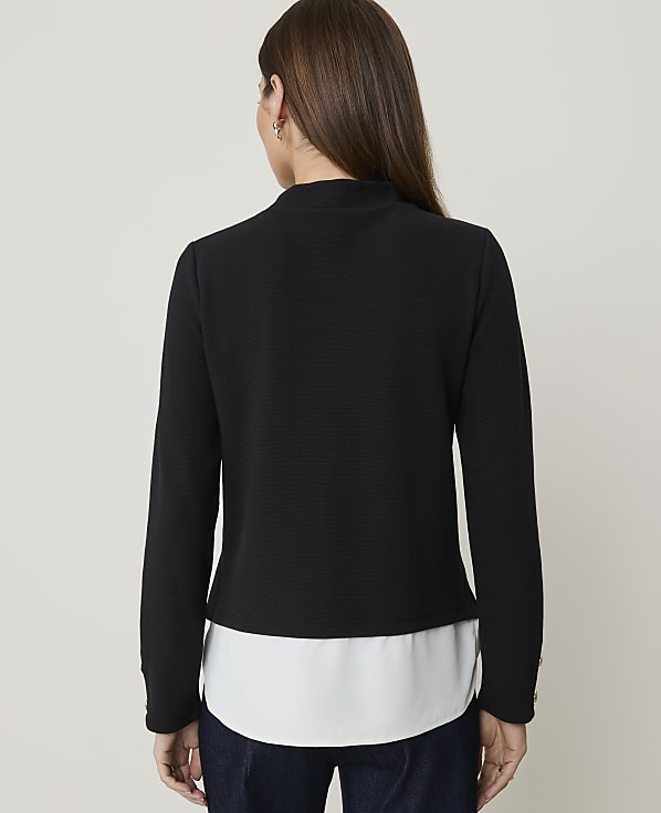 A woman in a black textured jacket over a white blouse stands against a plain, light-colored background.