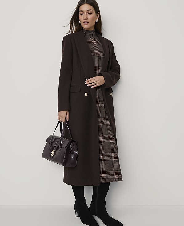A woman in a brown coat and plaid dress stands against a plain wall, holding a dark handbag.