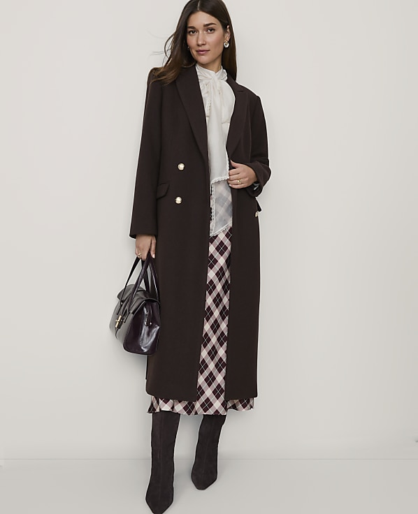 A woman in a long brown coat and plaid skirt stands against a plain white background, holding a dark handbag.