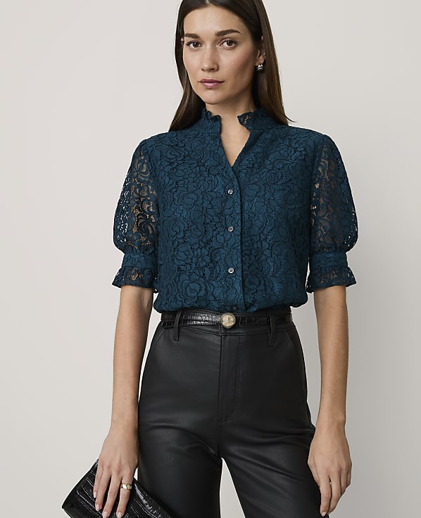 A woman in a teal lace blouse and black leather pants holds a black clutch in front of a plain white background.