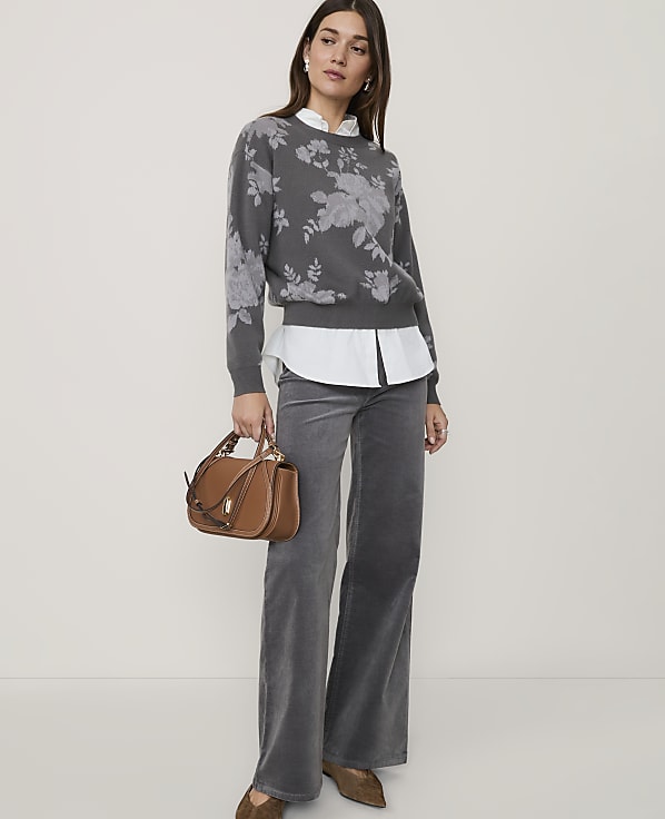 A woman in a floral sweater and gray pants stands with a brown handbag in front of a plain background.