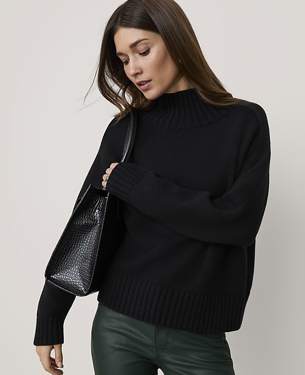 A woman in a black turtleneck sweater and green leather pants carries a black textured handbag against a plain gray background.