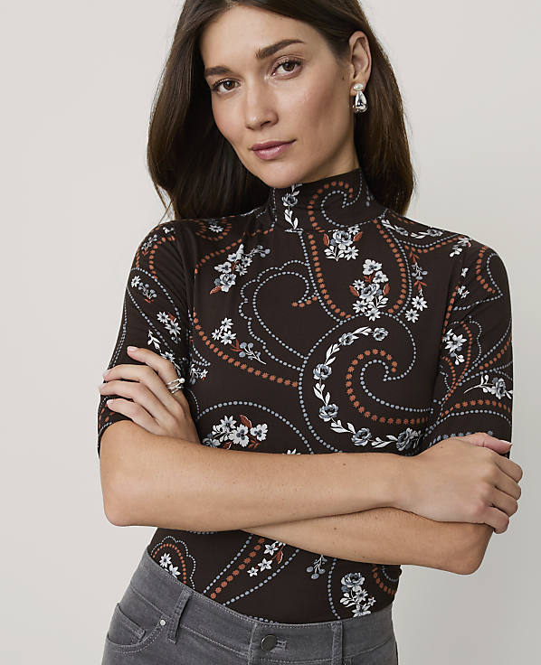 A woman with dark hair and a paisley-patterned turtleneck stands with her arms crossed against a plain background.