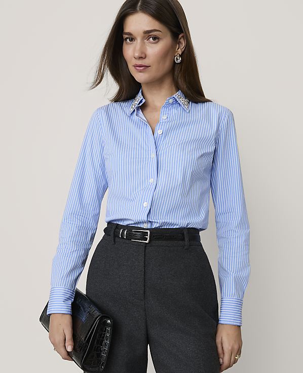 A woman in a blue pinstripe shirt and dark pants holds a black clutch in front of a plain background.