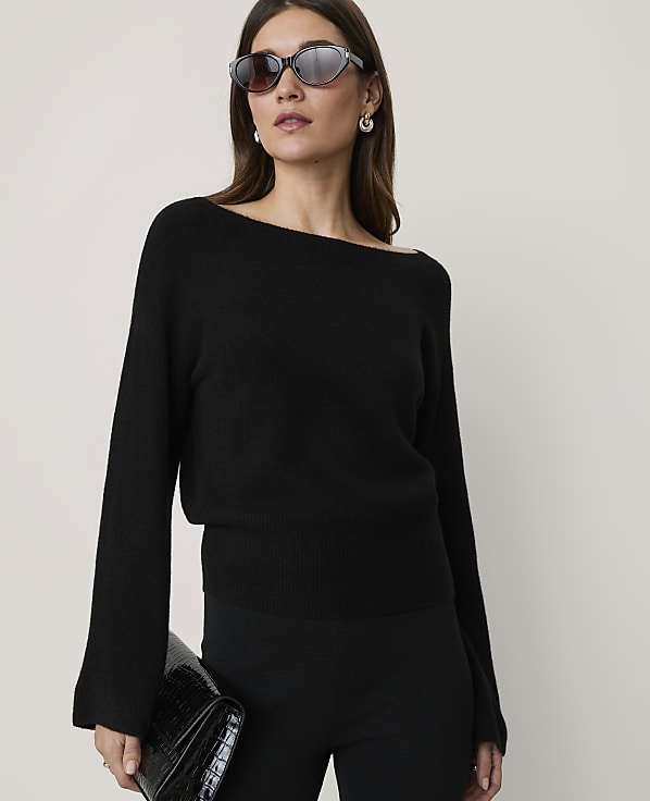 A woman in a black off-the-shoulder sweater and sunglasses holds a black clutch in front of a plain background.