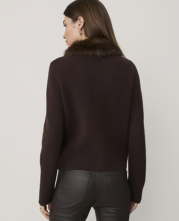 A woman in a brown fur-trimmed sweater and dark pants stands against a plain gray background.