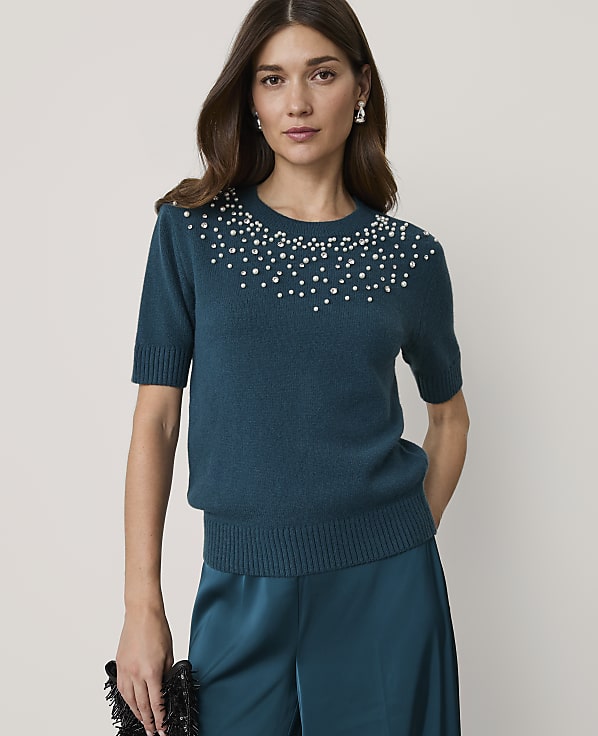 A woman in a teal sweater adorned with pearls stands against a plain, light background, holding a black clutch.