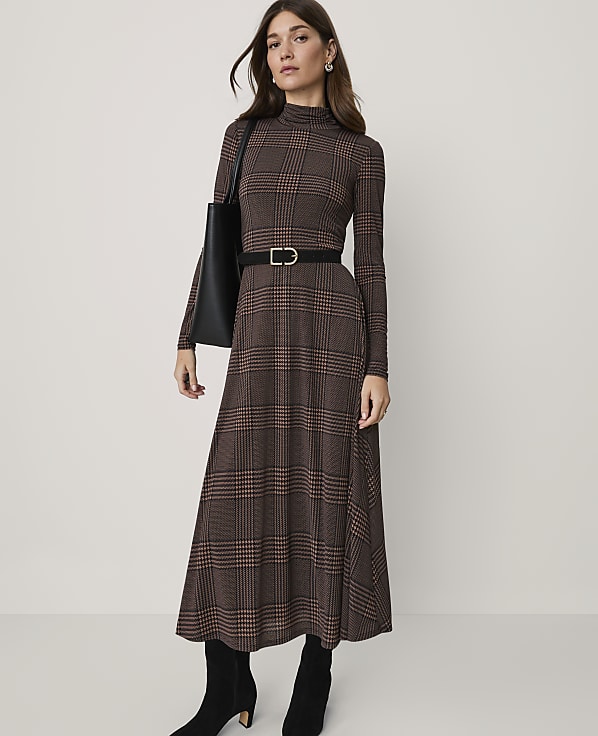 A woman in a brown plaid dress, black belt, and boots stands against a plain background.