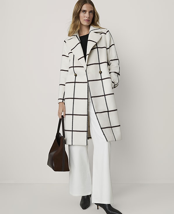 A woman in a white and black plaid coat and white pants stands in front of a plain white background, holding a brown purse.