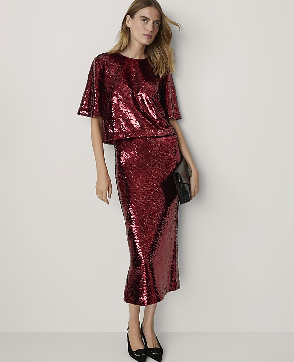 A woman in a sparkling red sequined top and skirt stands against a plain white background, holding a black clutch.