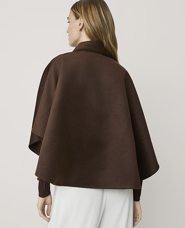 A woman from behind wears a brown cape over a maroon sweater and white pants against a plain background.