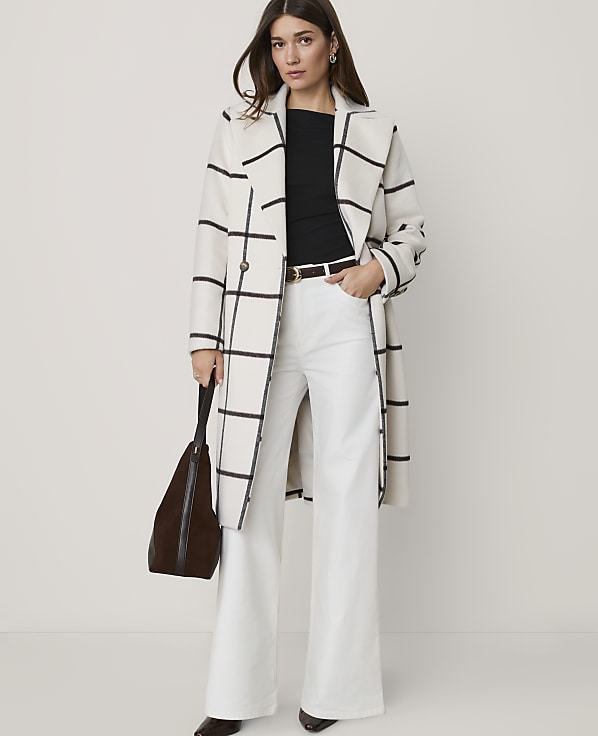 A woman in a cream and black checkered coat and white flared jeans stands against a plain white background, holding a brown bag.