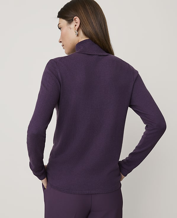 A woman in a purple turtleneck and pants stands against a plain, light-colored background.