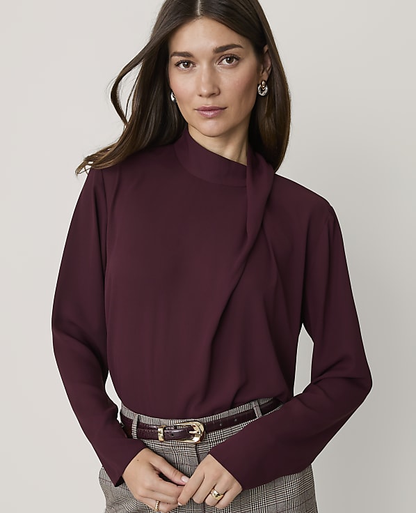 A woman in a burgundy blouse and plaid pants stands against a plain gray background.