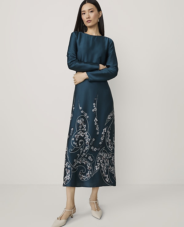 A woman in a teal dress with white floral embroidery stands with her arms crossed against a plain white background.