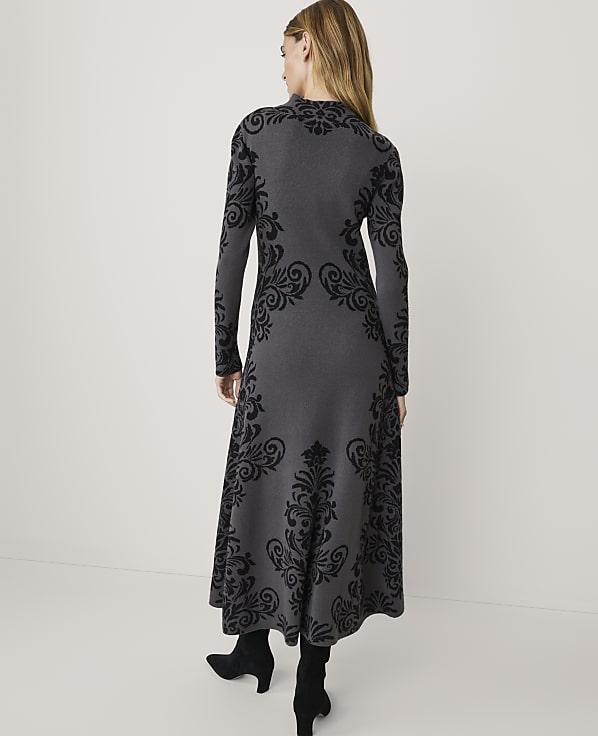 A woman in a long-sleeved gray dress with black ornate patterns stands against a plain white background.