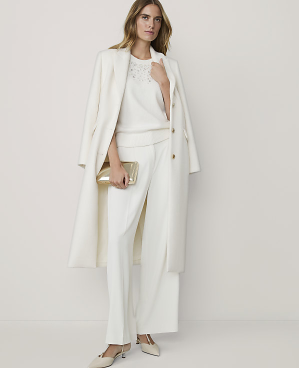 A woman in a white coat and matching pants holds a gold clutch in the foreground, against a plain white background.